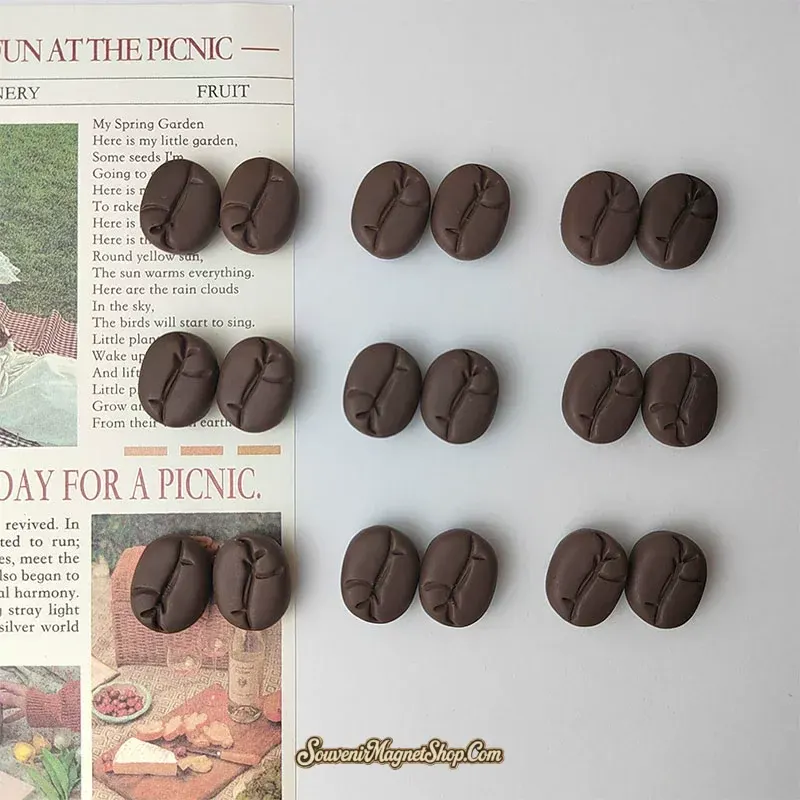 Coffee bean fridge magnets arranged in pairs across a white surface next to a vintage newspaper — set display showing multiple fun bean-shaped magnets and quantity