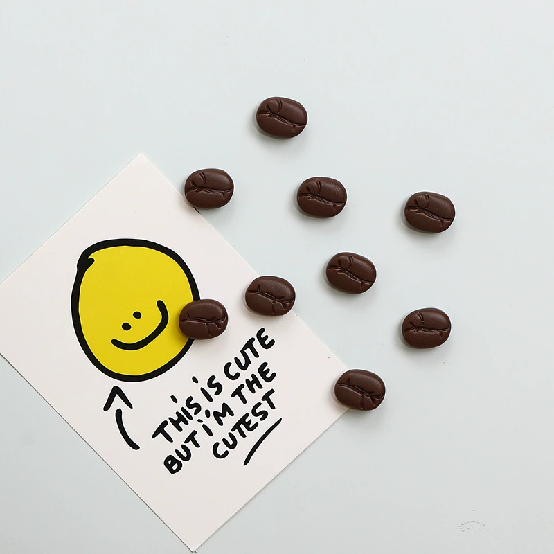 coffee-bean-fun-fridge-magnet-cutest-card Coffee bean fun fridge magnets scattered around a card with a bold yellow smiley face and handwritten text 'This Is Cute But I'm The Cutest' — dark brown bean-shaped magnets on pale blue background