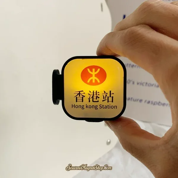 Hand holding lit Hong Kong Station MTR lightbox fridge magnet showing warm amber glow and orange logo
