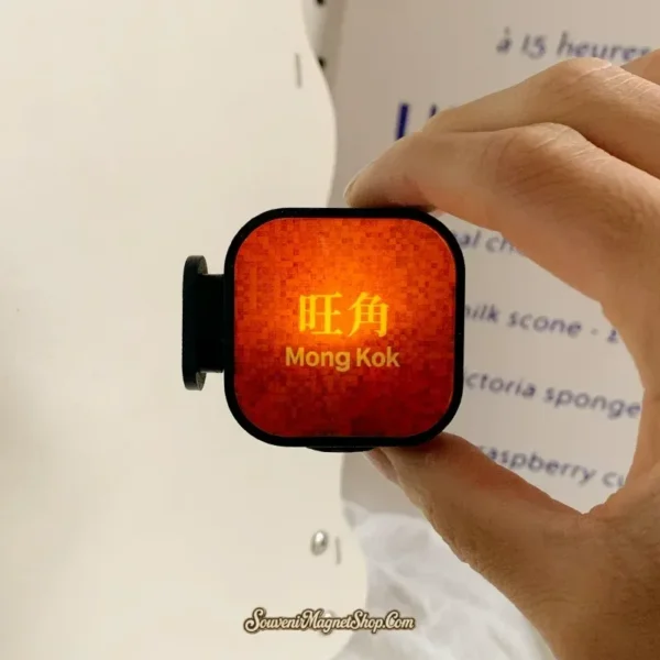 Hand holding lit Mong Kok MTR lightbox fridge magnet showing orange glow and gold Chinese text