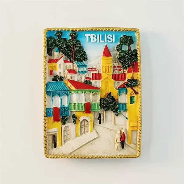 Georgia Tbilisi fridge magnet front view showing 3D old town street scene with yellow white buildings red tiled rooftops teal carved wooden balconies yellow church red conical spire miniature figures pale cobblestone lane TBILISI white text blue sky gold rope border frame