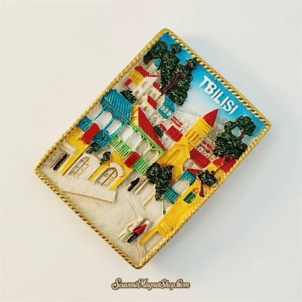 Tbilisi Georgia souvenir fridge magnet angled left view revealing 3D depth of gold rope framed resin magnet with carved wooden balconies church spire and colorful old town buildings