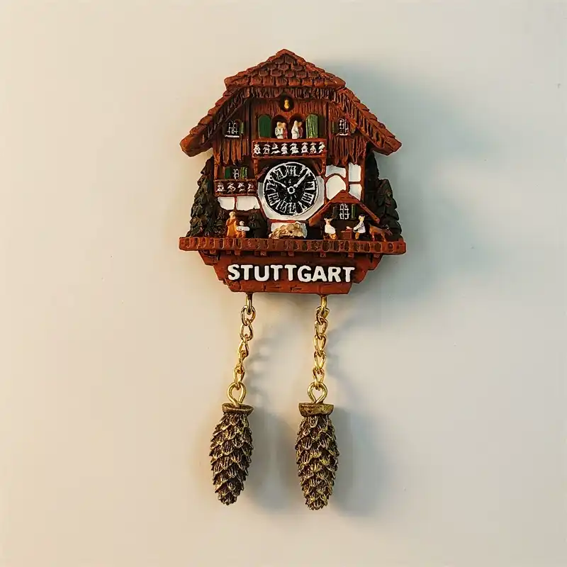 Germany Stuttgart fridge magnet front view showing 3D Black Forest cuckoo clock with dark brown carved chalet body black Roman numeral clock face green shuttered windows miniature figurines STUTTGART white lettering and two gold chains with bronze pine cone weights