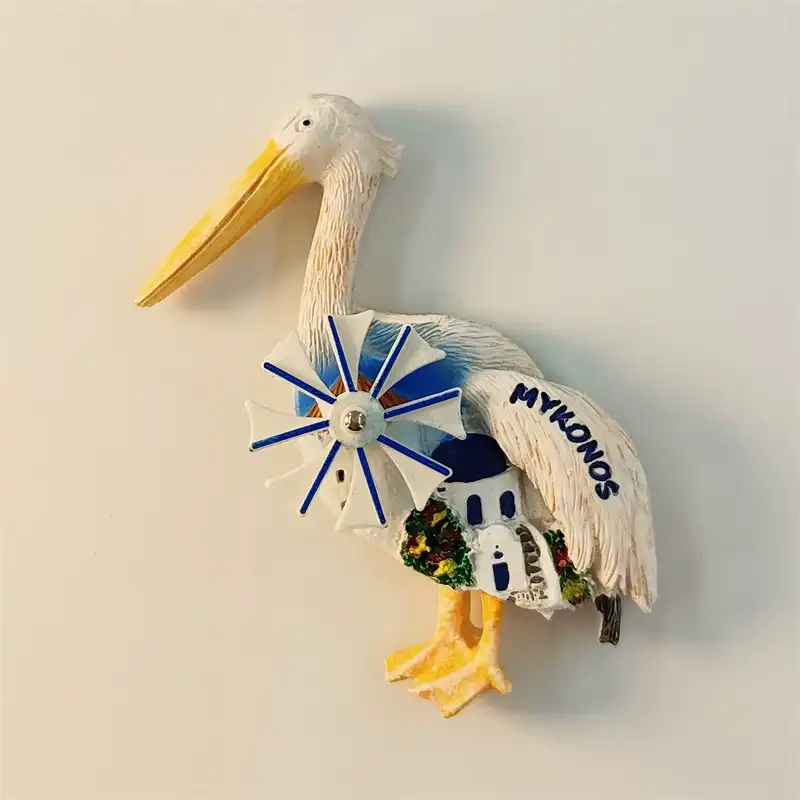 Greece Mykonos fridge magnet front view showing 3D white pelican figure with blue spinning windmill embedded in chest body miniature white blue Cycladic building green plants yellow beak webbed feet and MYKONOS blue lettering