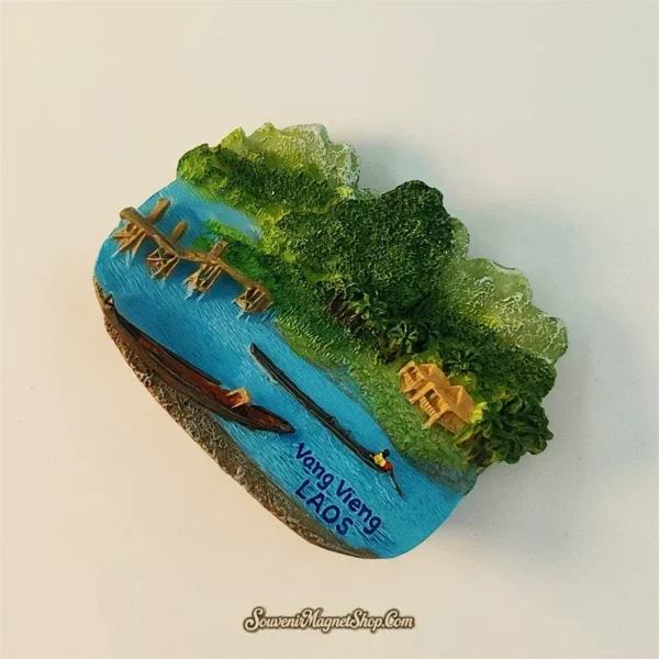 Laos Vang Vieng souvenir fridge magnet angled left view revealing 3D depth of bamboo bridge wooden boats and karst mountain relief above blue river