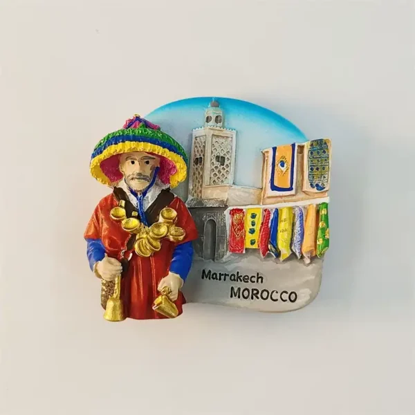 Front view of Morocco Marrakech water seller fridge magnet with colorful Guerrab figure, brass cups and Koutoubia Mosque background