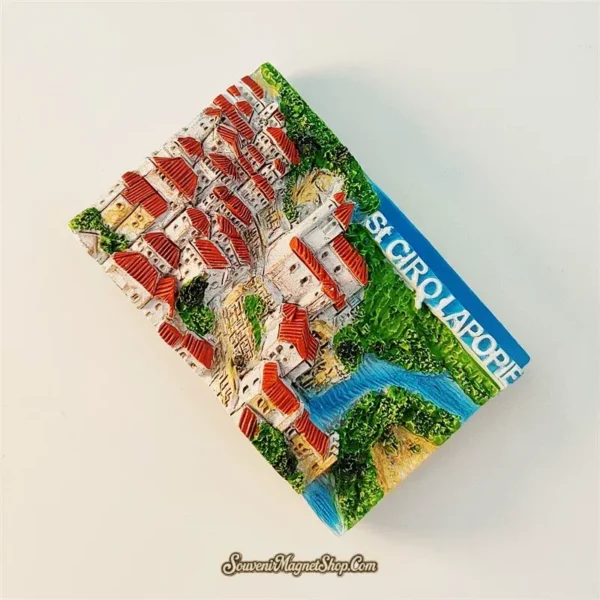 Angled view of hand-painted Saint-Cirq-Lapopie souvenir magnet showing raised 3D village relief