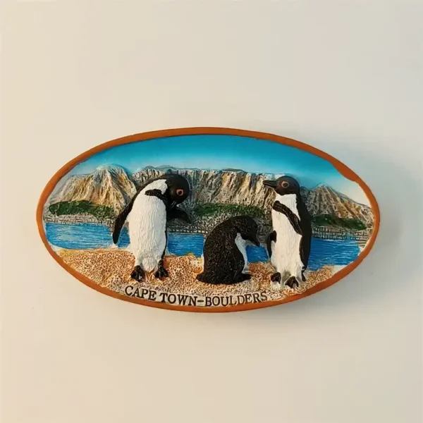 South Africa Cape Town fridge magnet front view showing 3D Boulders Beach scene with two adult African penguins flanking dark juvenile chick on sandy shore with turquoise ocean and Cape Peninsula mountains and Cape Town Boulders text