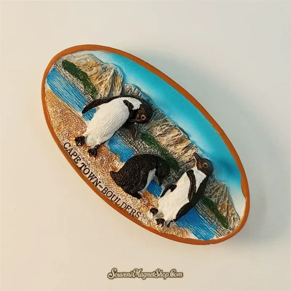 Cape Town Boulders Beach souvenir fridge magnet angled left view revealing 3D depth of African penguin figures sandy beach terracotta oval border and mountain backdrop detail