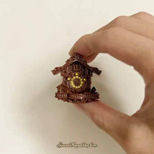 Hand holding the mini cuckoo clock magnet, showing the yellow dial, carved gables and pinecone weight