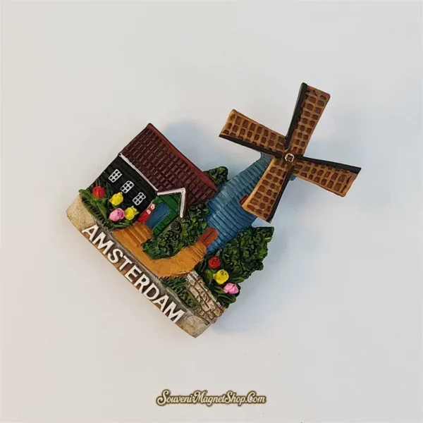 Amsterdam windmill magnet tilted clockwise, showing the lattice sails, blue mill tower and cottage garden above white AMSTERDAM lettering.
