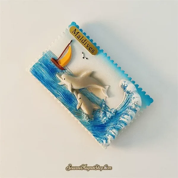 Angled view of the Maldives dolphin fridge magnet showing the raised dolphins, yellow sailboat, blue sea, and wave.