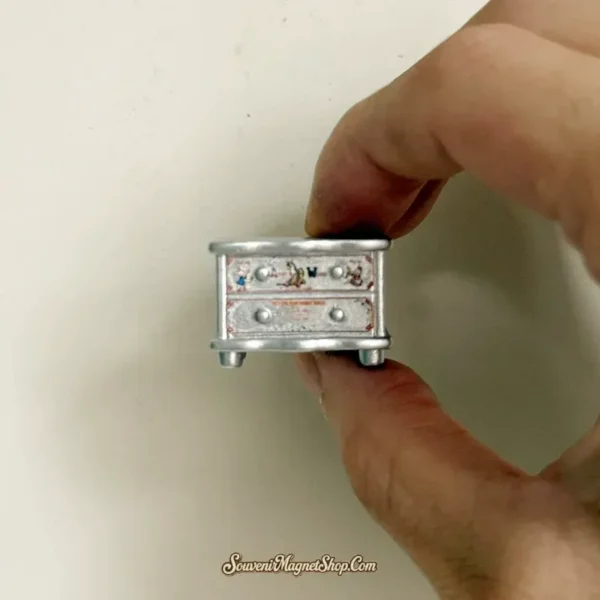 Cute silver bedside cabinet fridge magnet held between fingers, showing the raised top and two drawer fronts.