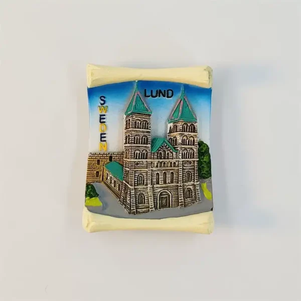 Front view of the Lund Cathedral fridge magnet in 3D relief, twin green-spired towers on tan sandstone with LUND and SWEDEN lettering, framed by a cream scroll border.