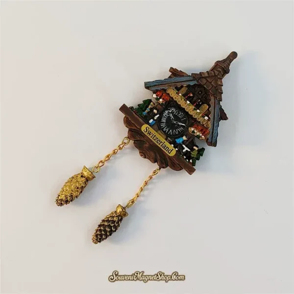 Switzerland flower-window cuckoo clock magnet tilted to the right, showing blue-grey eaves, a black dial and gold-tone chains with pinecone pendants.