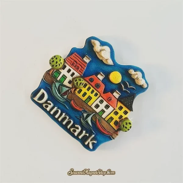 Denmark waterfront fridge magnet tilted clockwise, showing red-roofed houses, rounded green trees and boats on a blue background.