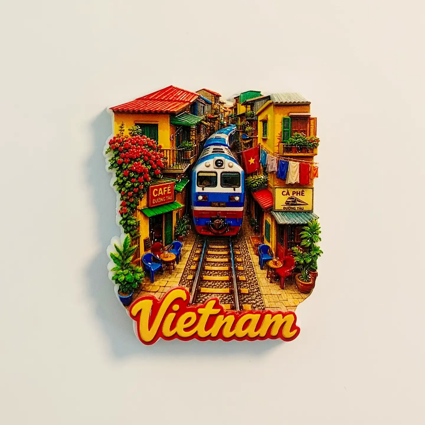 hanoi-train-street-fridge-magnet-front Hanoi Train Street fridge magnet with a blue locomotive between yellow café buildings, a Vietnamese flag and yellow Vietnam lettering.