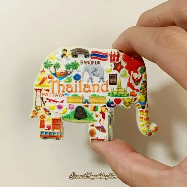 Thailand elephant-shaped fridge magnet held between fingers, showing orange lettering, a gray elephant and a travel icon collage.